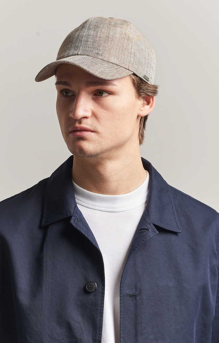A man wearing a textured cap, navy jacket, and white shirt looks to the side against a neutral background.
