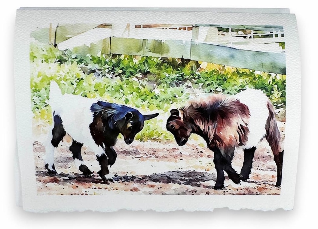 Watercolor-style card featuring two black-and-white goats facing each other on a dirt path, with soft greenery and a fence in the background, printed on textured paper with deckled edges.