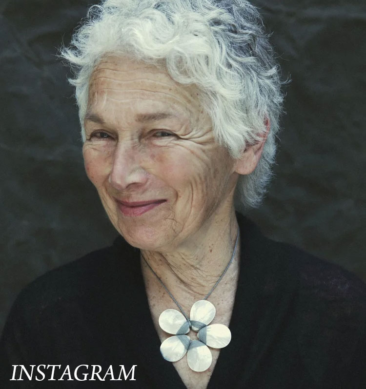 Close-up portrait of an older woman with short, curly white hair and a warm, subtle smile. She wears a simple dark top and a sculptural, light-toned statement necklace, set against a softly textured dark background.