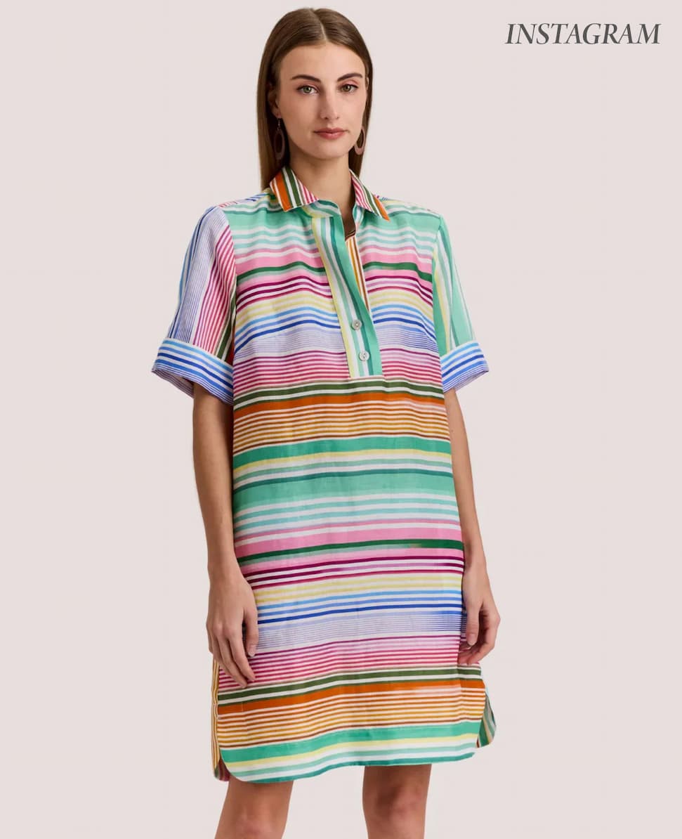 A woman models a relaxed, short-sleeve shirt dress in colorful horizontal stripes, styled with simple earrings against a neutral background.