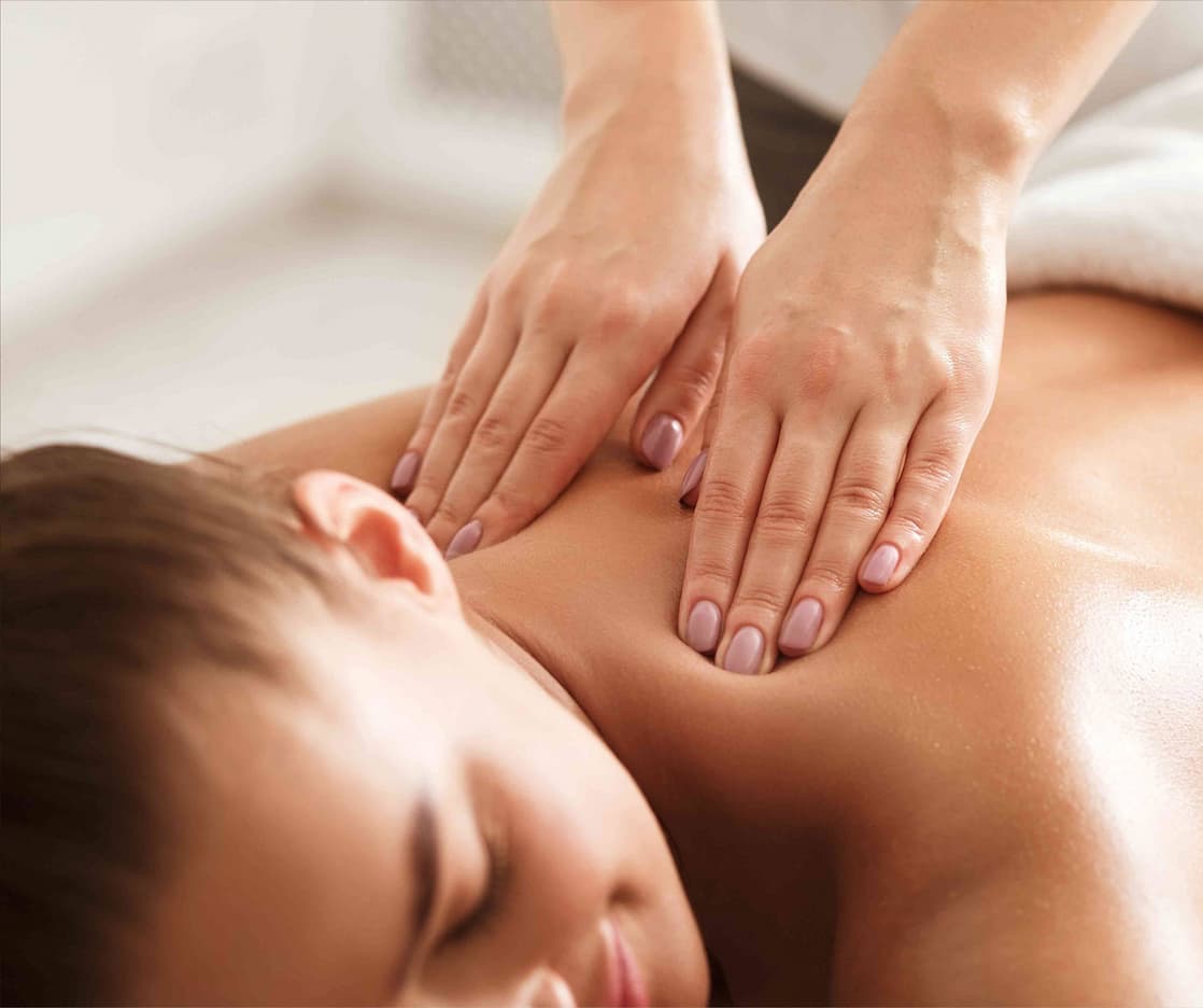 A person is lying face down receiving a back massage. The masseuse’s hands are gently pressing on the person’s upper back and shoulders. The setting appears calm and relaxing. Fearrington Village