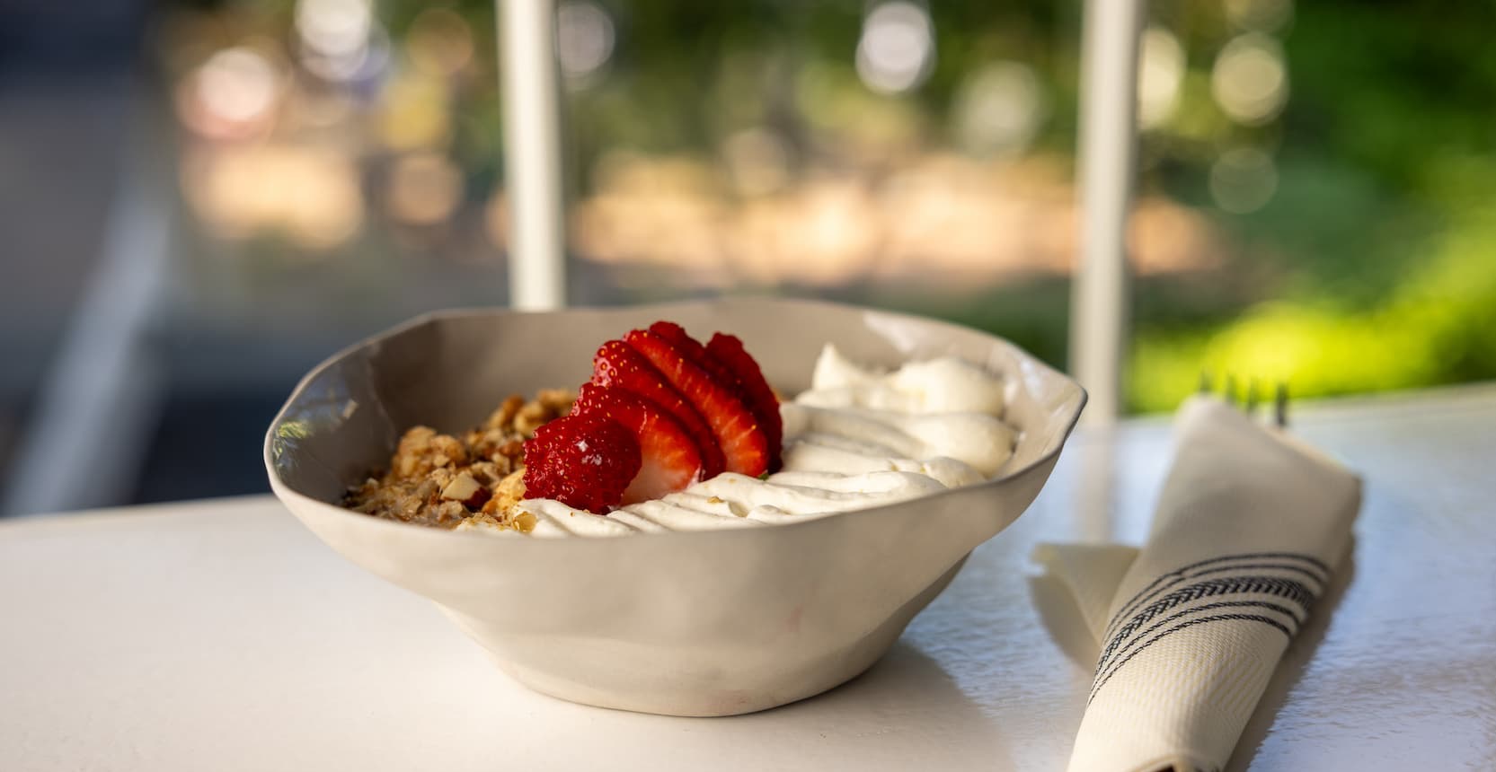 Stoneware bowl filled with creamy yogurt topped with sliced strawberries, granola, and swirls of whipped cream. Set on a white tabletop beside a striped linen napkin, the dish is softly lit by natural light from a nearby window overlooking lush greenery.