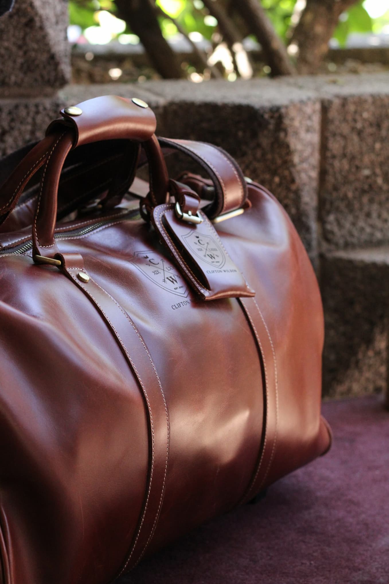Close-up of a rich brown leather duffel bag with stitched detailing and brass hardware, resting outdoors against a stone backdrop with greenery.
