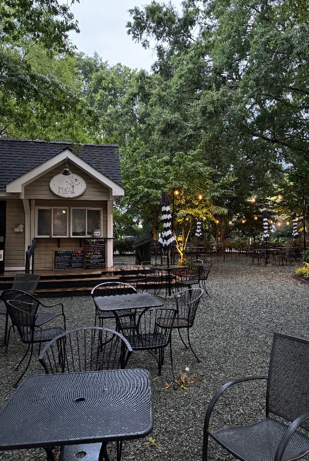 A quaint outdoor café with a small wooden building featuring a menu board and sign that reads "Food". The seating area has wrought-iron tables and chairs on a gravel ground. Black-and-white striped umbrellas are scattered around. Tall green trees surround the serene setting. Fearrington Village