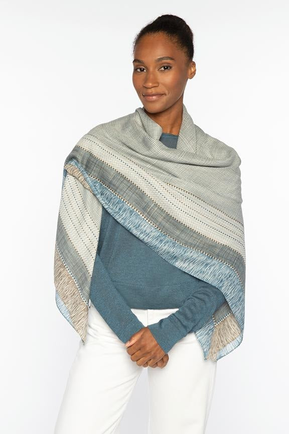 Woman wearing a soft blue sweater and white pants, styled with a textured striped shawl in muted green, blue, and cream tones draped over her shoulders.