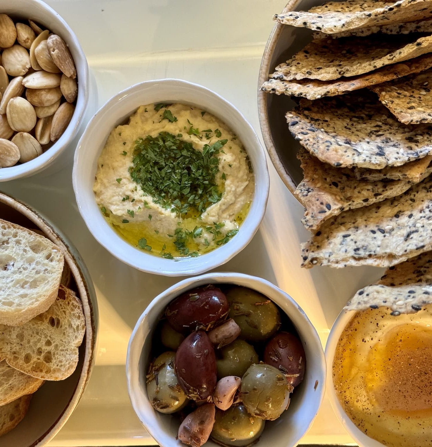 A beautifully arranged appetizer board featuring a bowl of creamy hummus topped with olive oil and fresh herbs, surrounded by seeded crackers, marinated olives and garlic, toasted bread slices, and roasted pumpkin seeds. Warm natural light highlights the textures and colors, creating an inviting spread perfect for sharing.