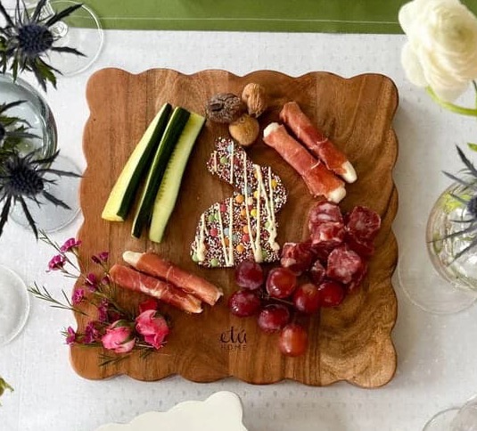 Overhead view of a scalloped acacia wood serving board arranged with a charcuterie spread featuring grapes, salami, prosciutto-wrapped cheese sticks, cucumber spears, nuts, and a decorated wedge of cheese. Fresh flowers and glass vases frame the board, creating an elegant entertaining scene.