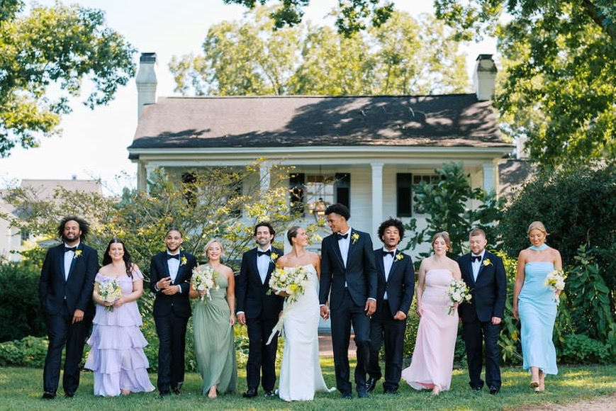 Wedding party walking across the lawn in front of a white farmhouse at Fearrington Village, with the bride and groom centered and attendants in formal attire carrying bouquets.