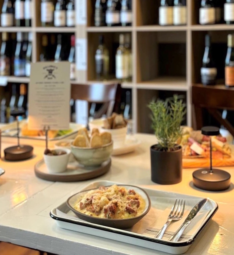 A cozy wine bar table set with a creamy pasta dish topped with grated cheese, served on a tray with silverware. In the softly blurred background, shelves of wine bottles, small table lamps, bread and charcuterie plates, and a potted herb create a warm, intimate dining atmosphere.