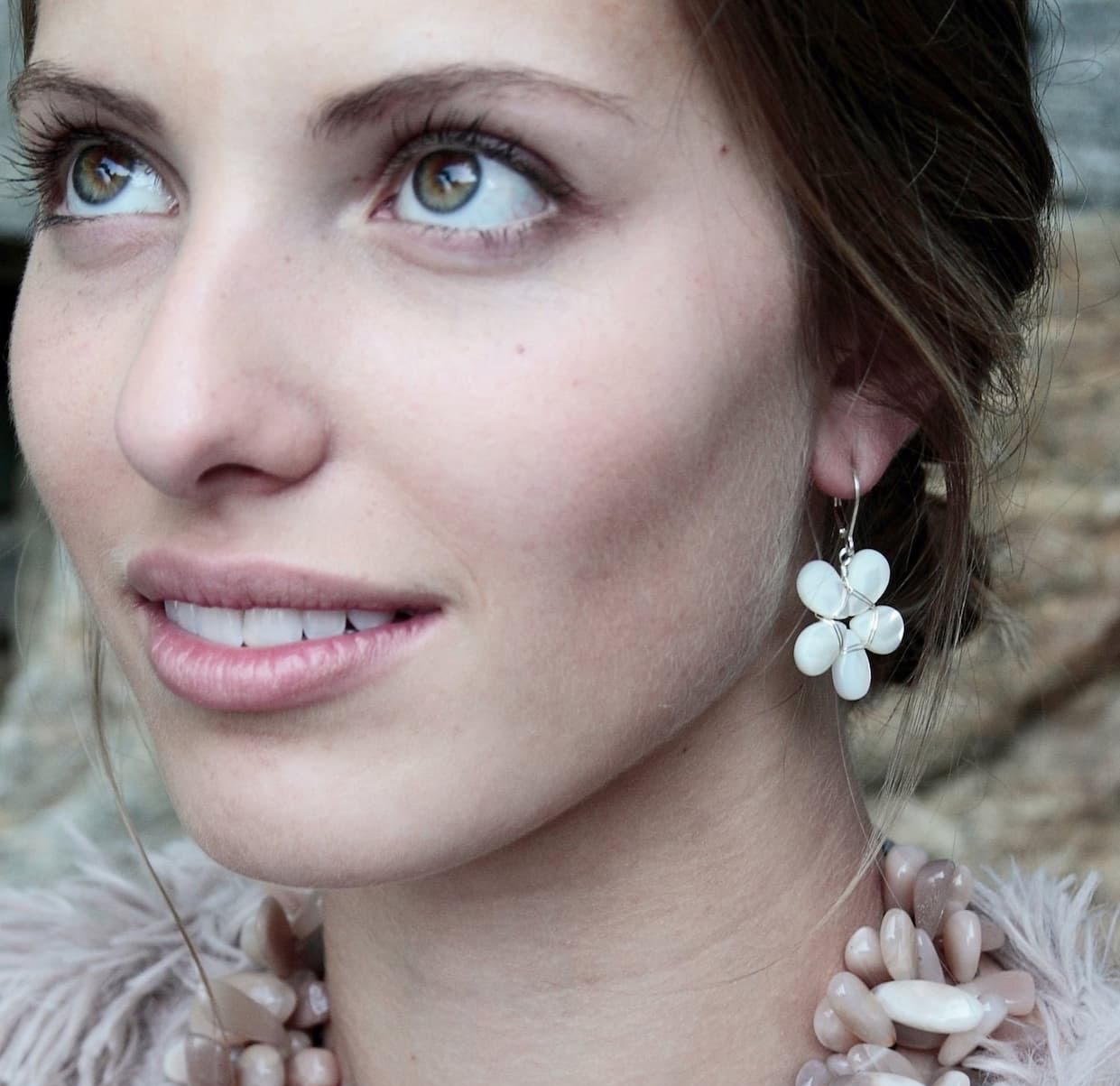 Close-up portrait of a woman with soft, natural makeup and green eyes, looking slightly upward. She wears a white floral drop earring and a chunky beaded necklace in neutral tones, with a softly blurred outdoor background.