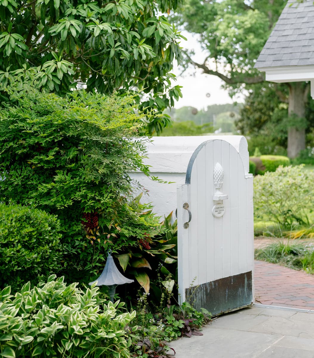 White arched garden gate framed by lush greenery, opening onto a brick pathway that leads into a peaceful courtyard. Layered shrubs, hostas, and mature trees create a secluded, welcoming entrance with classic Southern garden charm. Fearrington Village.