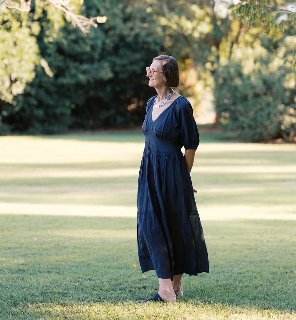 Gilda McDaniel wearing glasses and a navy blue dress stands barefoot on a grassy lawn, looking to the side beneath leafy trees in soft afternoon light.