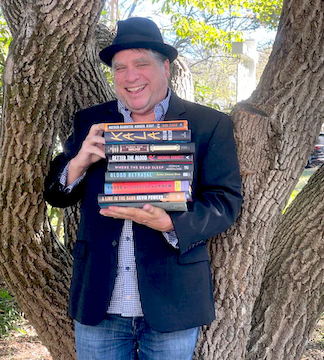 Pete Mock wearing a black hat and jacket stands in front of a tree, smiling and holding a stack of books labeled as Beltie Mystery Prize nominees for 2026.