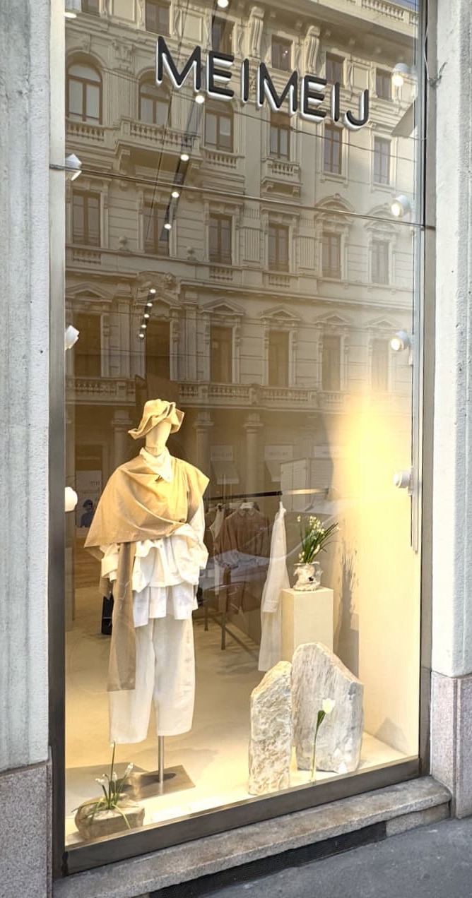 Boutique storefront window with a mannequin dressed in layered neutral clothing and hat, styled among stone accents and white flowers, with a city building reflected in the glass.