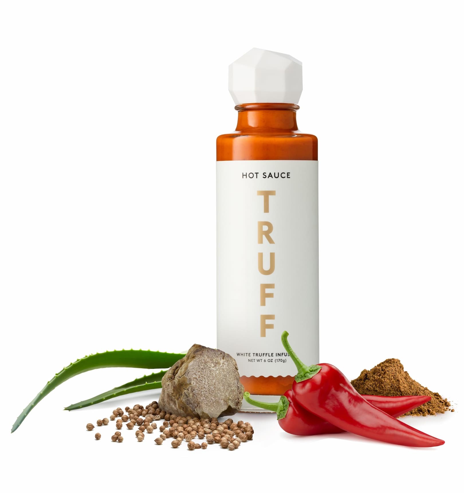 Bottle of TRUFF white truffle-infused hot sauce with a faceted white cap, styled alongside fresh red chili peppers, a truffle, coriander seeds, spices, and an aloe leaf on a clean white background. The arrangement highlights the sauce’s premium ingredients and gourmet appeal.