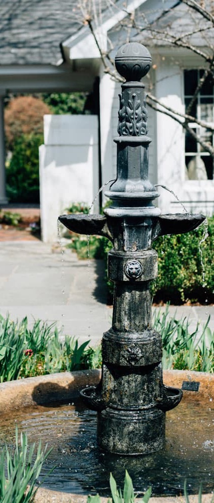 Ornate black metal fountain with multiple tiers gently spilling water into a shallow basin, set in a landscaped garden near a white building.