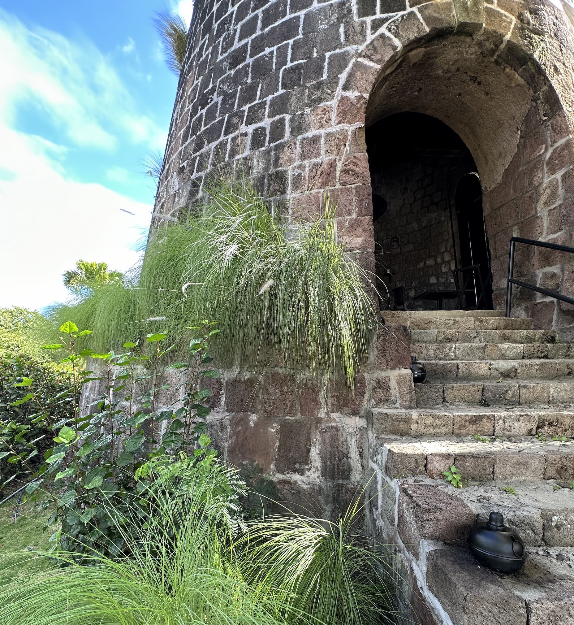 A stone tower with an arched doorway and curved steps, surrounded by tropical grasses and plants under a bright blue sky in Nevis.