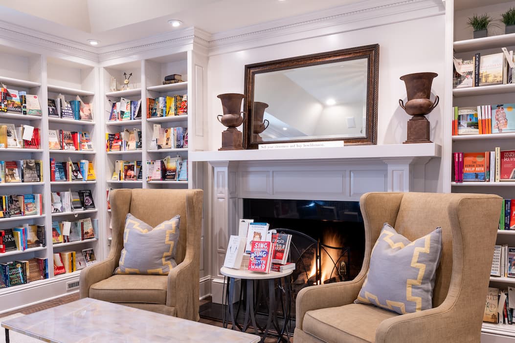 Cozy reading nook inside a bookstore with two upholstered armchairs facing a lit fireplace, a small table displaying books between them, and tall white bookshelves filled with colorful titles lining the walls.