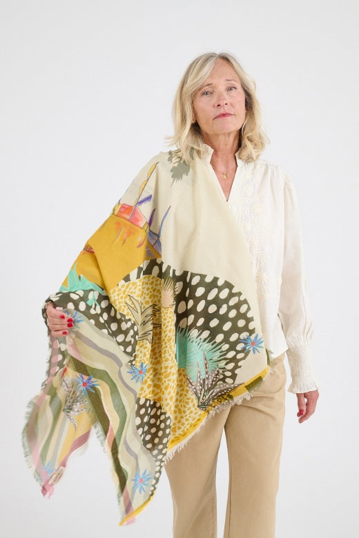 Woman wearing a cream blouse and neutral pants, styled with a large, flowing scarf in soft yellow, green, and patterned prints draped over one shoulder.
