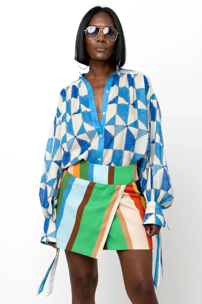 A woman stands against a white background wearing oversized sunglasses, a loose blue-and-cream geometric print blouse, and a high-waisted wrap skirt with bold vertical stripes in green, blue, tan, and orange. Her hair is styled in a sleek bob, and the look feels modern and graphic.