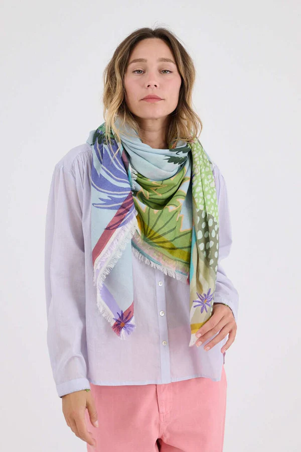 Woman wearing a light button-down shirt and pink pants, styled with a large, colorful patterned scarf draped around her neck against a neutral background.