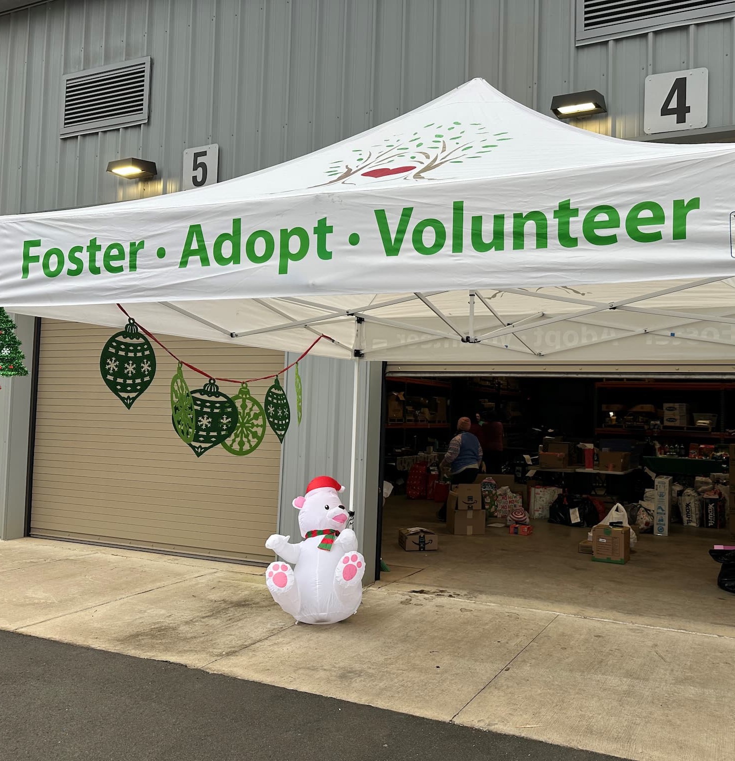 White canopy tent outside a warehouse-style building with a banner reading “Foster · Adopt · Volunteer,” decorated with green holiday ornaments and a small inflatable polar bear, with boxes and volunteers visible inside the open garage.