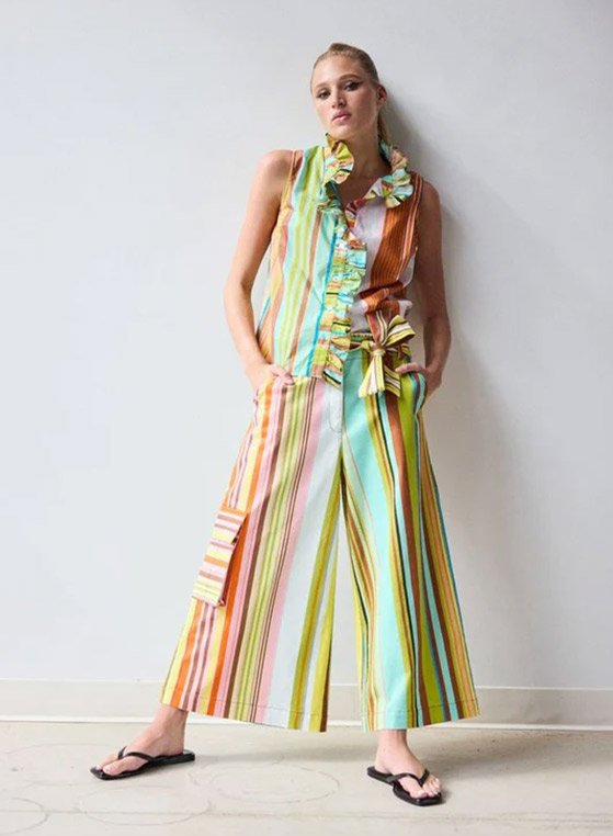 Woman wearing a sleeveless striped blouse with ruffle detailing and matching wide-leg cropped pants, featuring colorful vertical stripes in aqua, coral, yellow, pink, and olive. The coordinated set is finished with a striped waist tie and styled with black flip-flops against a neutral backdrop.