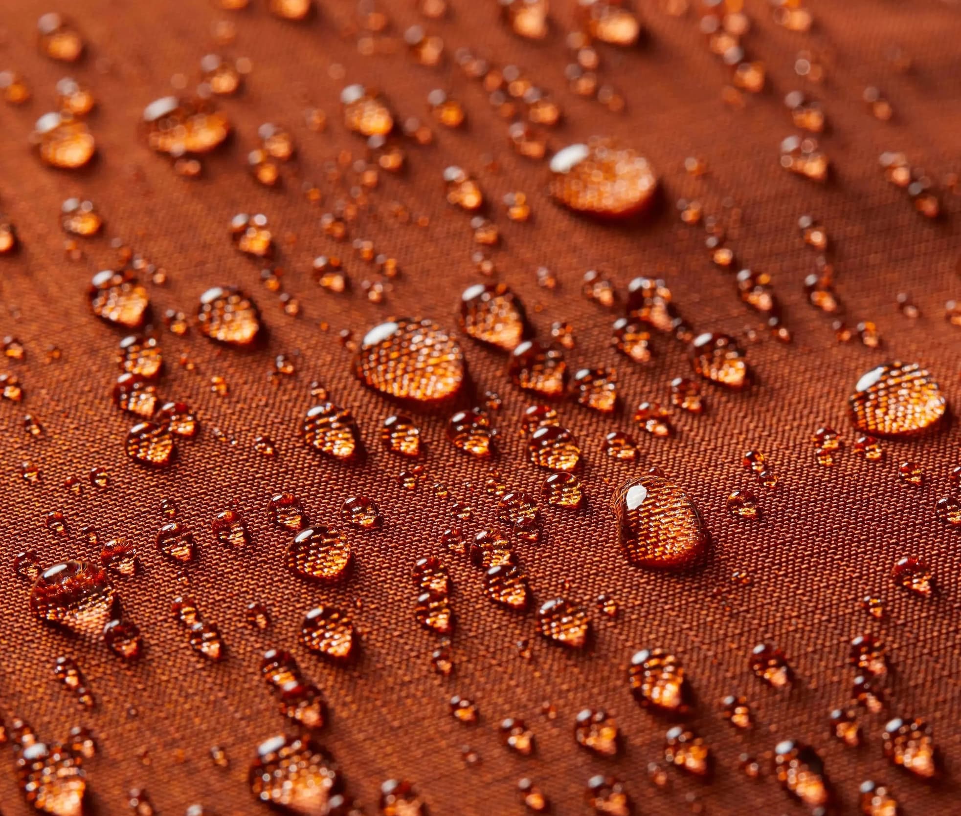 A close-up macro image of water droplets beading on rust-colored technical fabric. The droplets sit on the surface without soaking in, highlighting the material’s water-resistant finish.