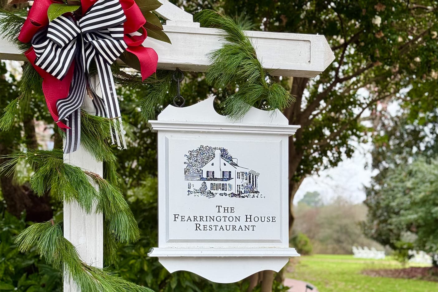 A white sign with an illustration of a house reads “The Fearrington House Restaurant.” The sign hangs from a post decorated with pine garland and a black, white, and red bow, with trees and greenery in the background. Fearrington Village