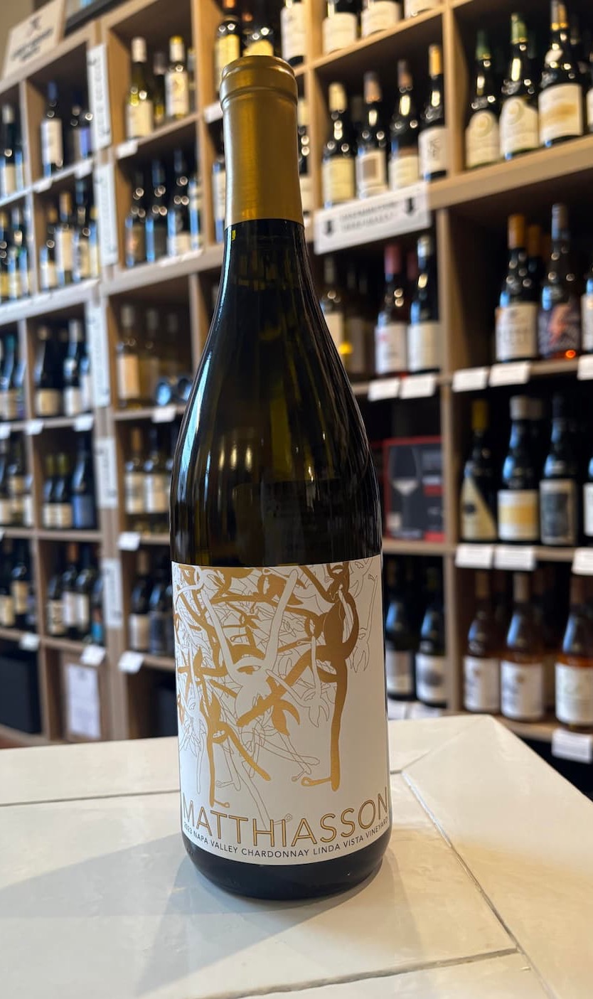 A bottle of Matthiasson Linda Vista Vineyard Chardonnay stands on a white countertop in front of a wall of wine shelves. The bottle features a minimalist cream-and-gold label with abstract vine illustrations. Behind it, rows of wine bottles softly blur into the background, creating a warm, inviting wine shop atmosphere.