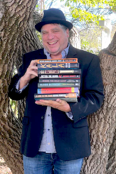 Pete Mock standing under a tree holding the 8 Beltie Mystery Prize longlist titles for 2026.