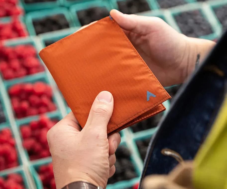 A pair of hands holds a compact orange travel wallet with a small blue logo in the corner. In the blurred background are baskets of fresh berries at an outdoor market.