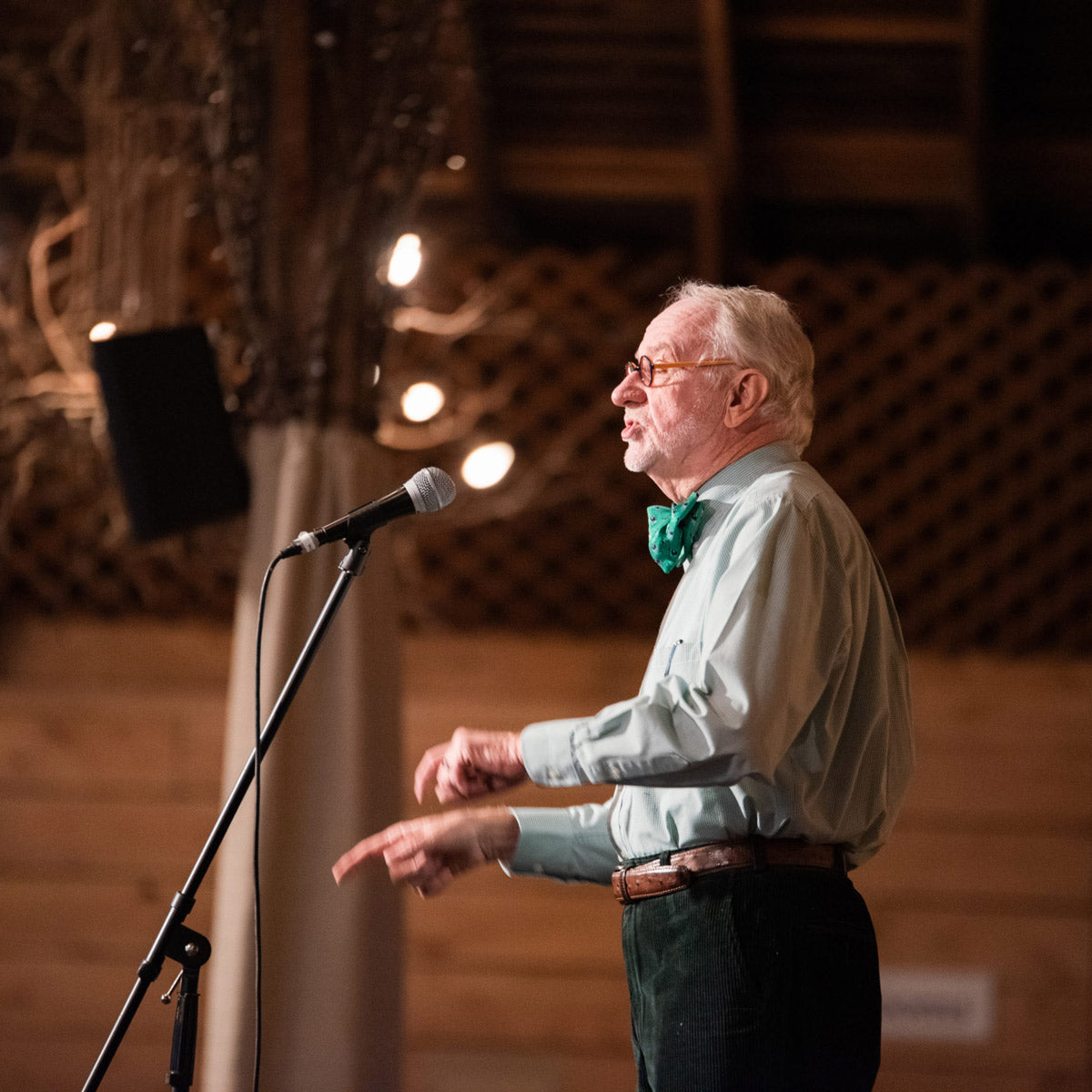An elderly man with white hair and glasses, wearing a green bow tie and light shirt, speaks animatedly into a microphone on a stand in a warmly lit indoor setting. Fearrington Village