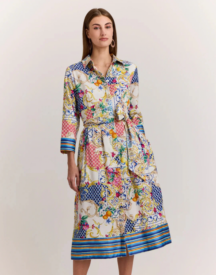 A woman models a long-sleeve shirt dress in a vibrant mixed print with florals, tiles, and stripes, finished with a tie waist and statement earrings.