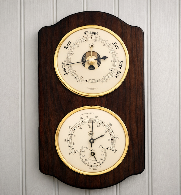 Wall-mounted dual-dial barometer and hygrometer in a dark wood case with brass bezels, displayed on light gray painted beadboard paneling.