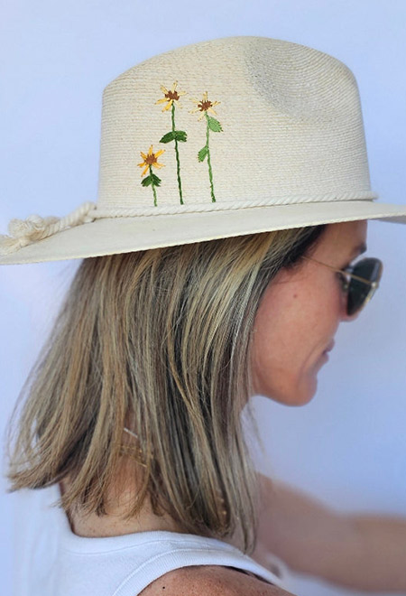 Side profile of a woman wearing an ivory handwoven straw hat embroidered with three delicate yellow wildflowers, paired with sunglasses and a white sleeveless top. Fearrington Village.