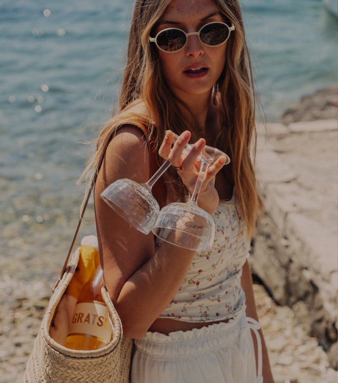 Woman at the beach with a bag containing a bottle of Gratsi Sparkling Wine, holding 2 champagne glasses by the stems. 