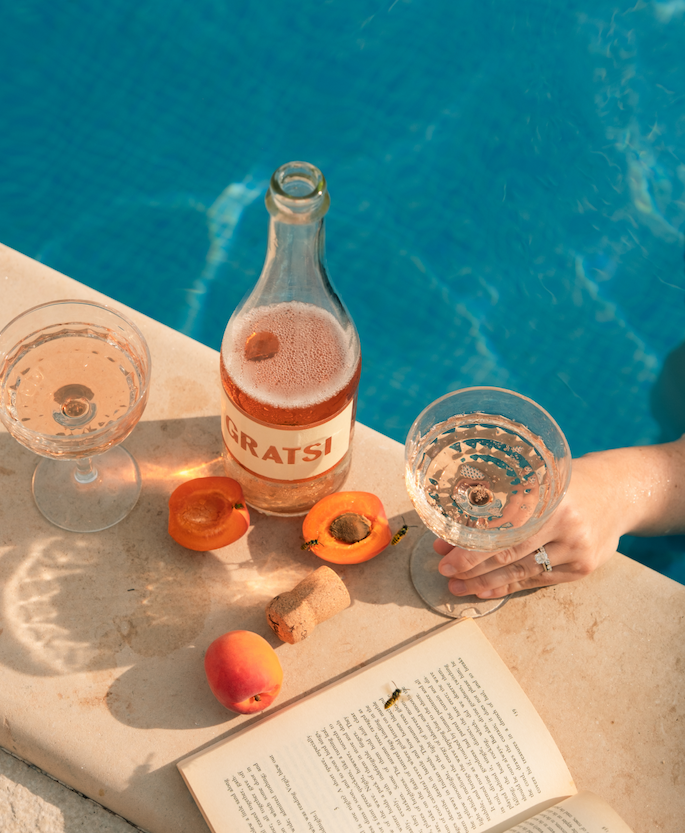 Poolside with a bottle of Gratsi Sparkling Rosé, a book, fruit and 2 glasses.