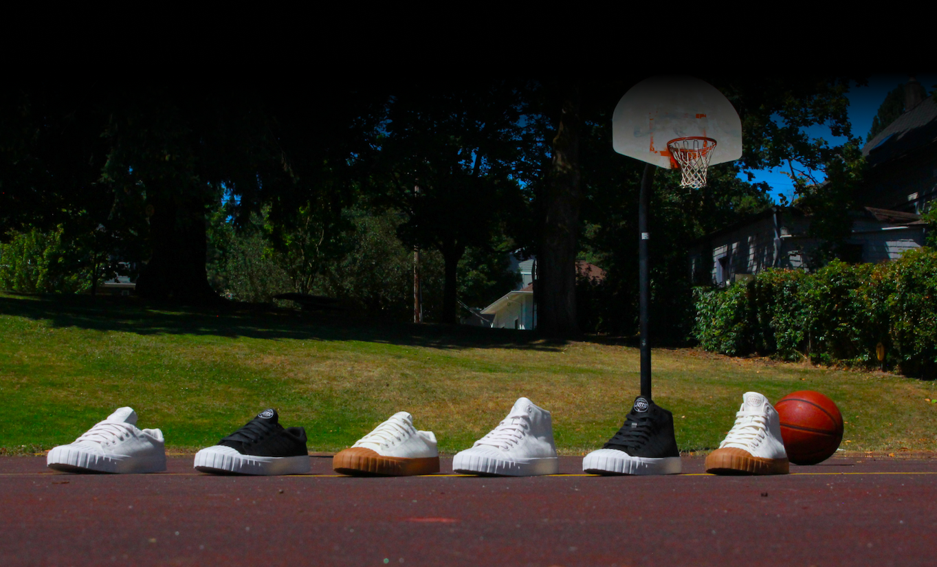A row of six Legacy sneakers lined up on a park basketball court beside a basketball, with a hoop and backboard behind them.