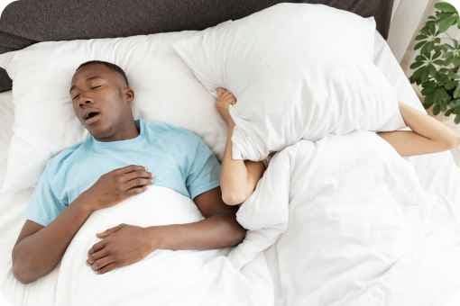 Man snoring in bed, partner has a pillow over their face to muffle the sound