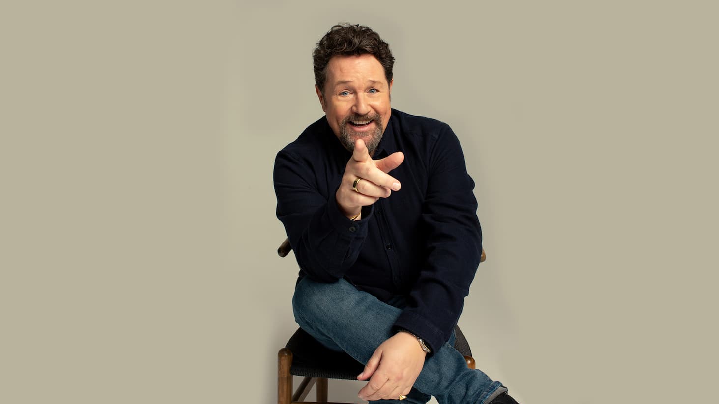 Michael Ball seated on a chair with legs crossed, pointing toward the camera, wearing a dark shirt and jeans against a plain light background.