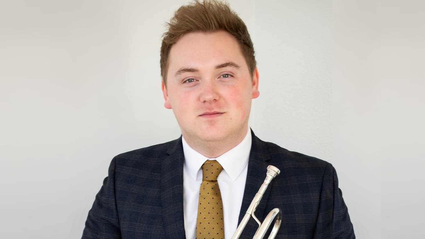 Professional portrait of a musician in a suit holding a silver trumpet against a light background, facing the camera in a formal promotional photograph.
