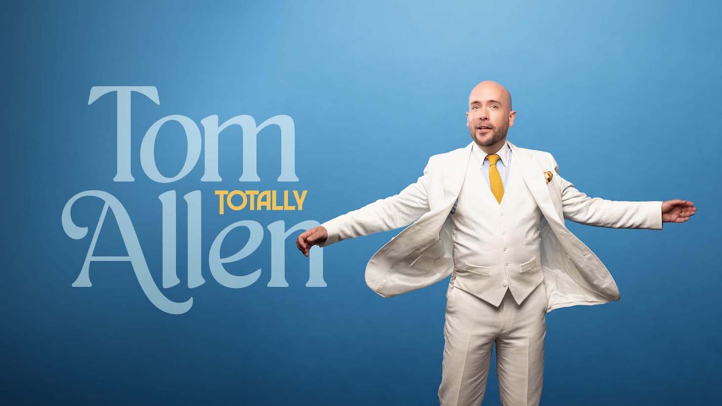 Tom Allen in a white suit with yellow tie stands arms outstretched against blue background with large text reading "Tom Totally Allen".
