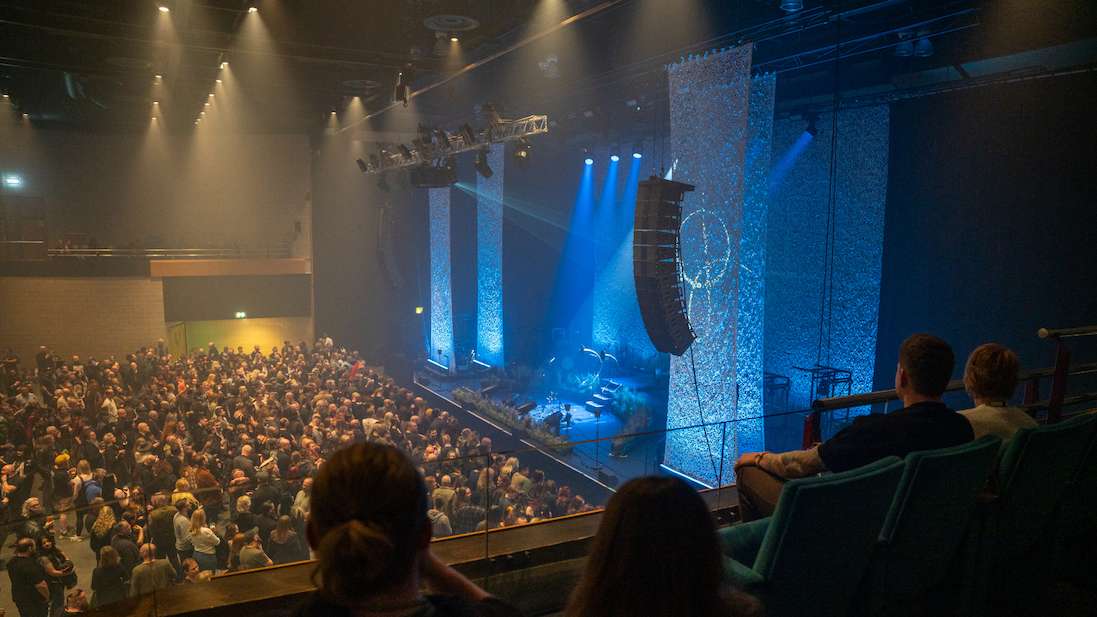Wide view of York Barbican from a balcony, showing a large standing crowd gathered in front of a stage. The stage is lit with blue spotlights and framed by tall, textured panels, with speakers and lighting rigs hanging above. In the foreground, several seated audience members are visible in dim light, while the main floor is densely packed with people waiting for the performance.