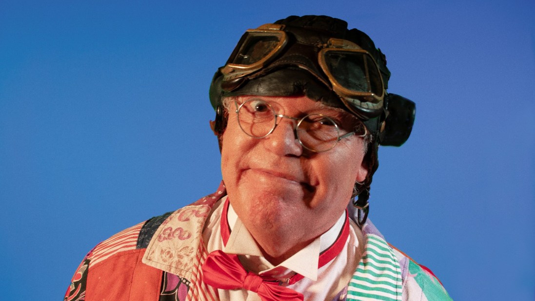 Close-up portrait of Chubby wearing round glasses and vintage-style aviator goggles on their head, set against a plain blue background. Chubs is dressed in a colourful patterned shirt, red bow tie, and suspenders, facing the camera with a slight smile.