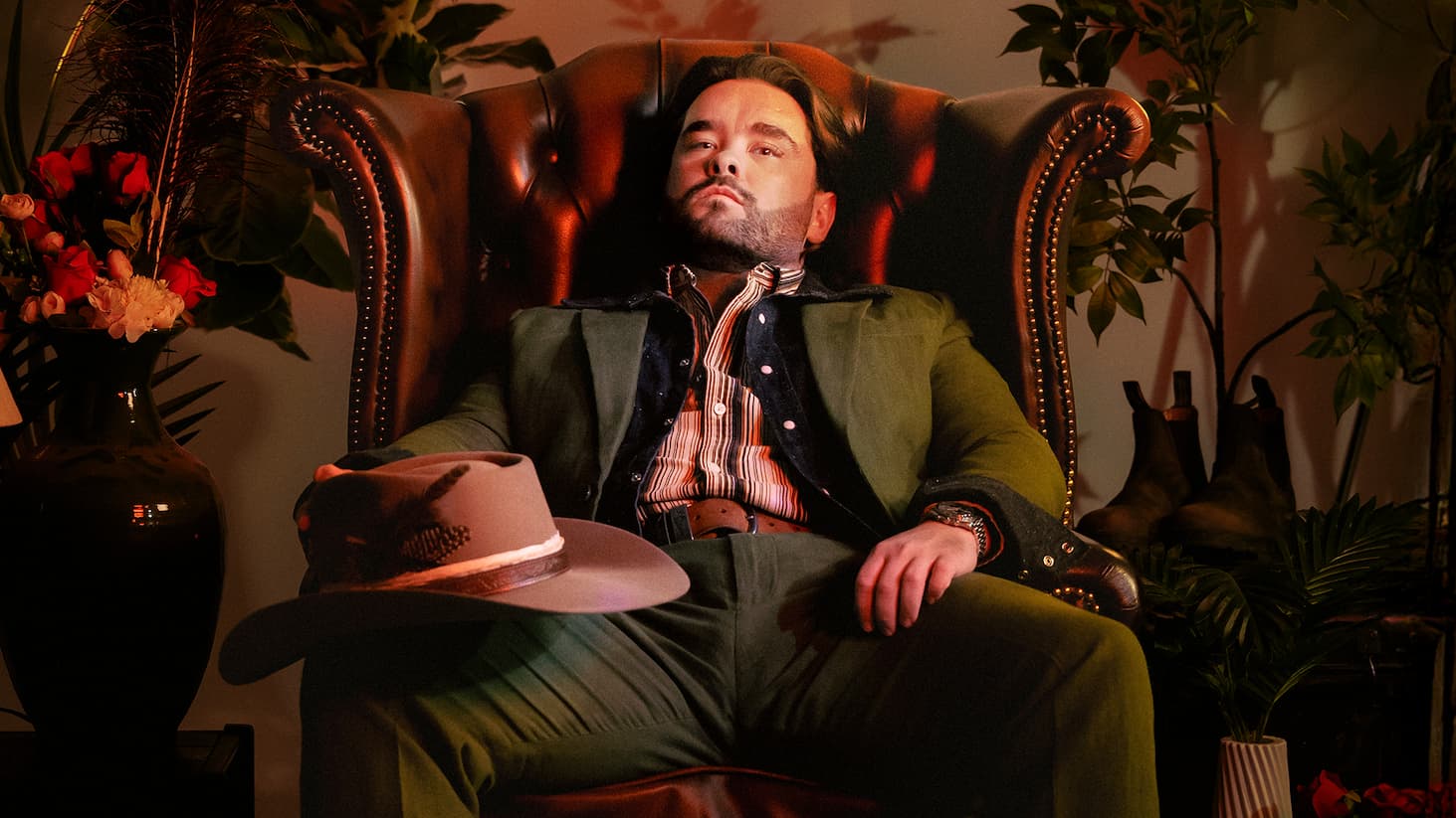 Studio portrait of Adam Rowe reclining in a brown leather armchair, wearing a green suit and layered shirt. A wide-brim hat rests on the chair arm in the foreground. The setting includes warm, low lighting, leafy plants, a dark vase with flowers on the left, and boots on the floor to the right, creating a moody, styled interior scene.