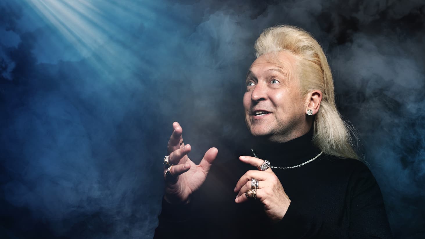 Studio portrait of Clinton with long, slicked-back hair wearing a black turtleneck, multiple rings, and a chain necklace, set against a smoky dark blue background. Dramatic beams of light shine from the upper left, illuminating the scene as Clinton raises both hands mid-gesture.