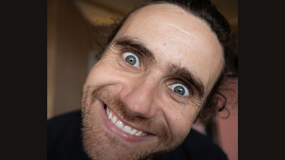 Close-up of Tom Lawrinson leaning toward the camera with a broad smile, wide eyes and blurred indoor background.
