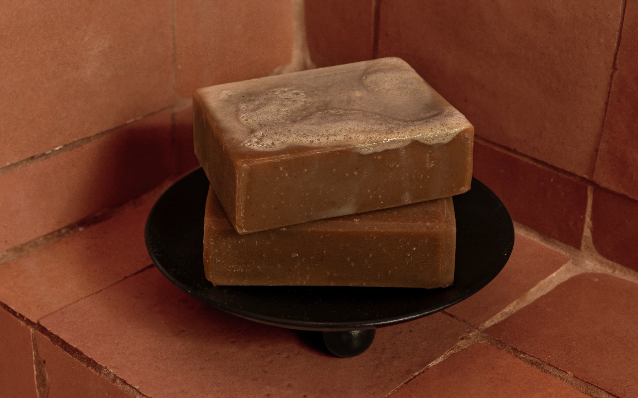 A soap dish set against square, terracotta tiles, stacked with two bars of Black soap - the top bar coated with sudsy foam.