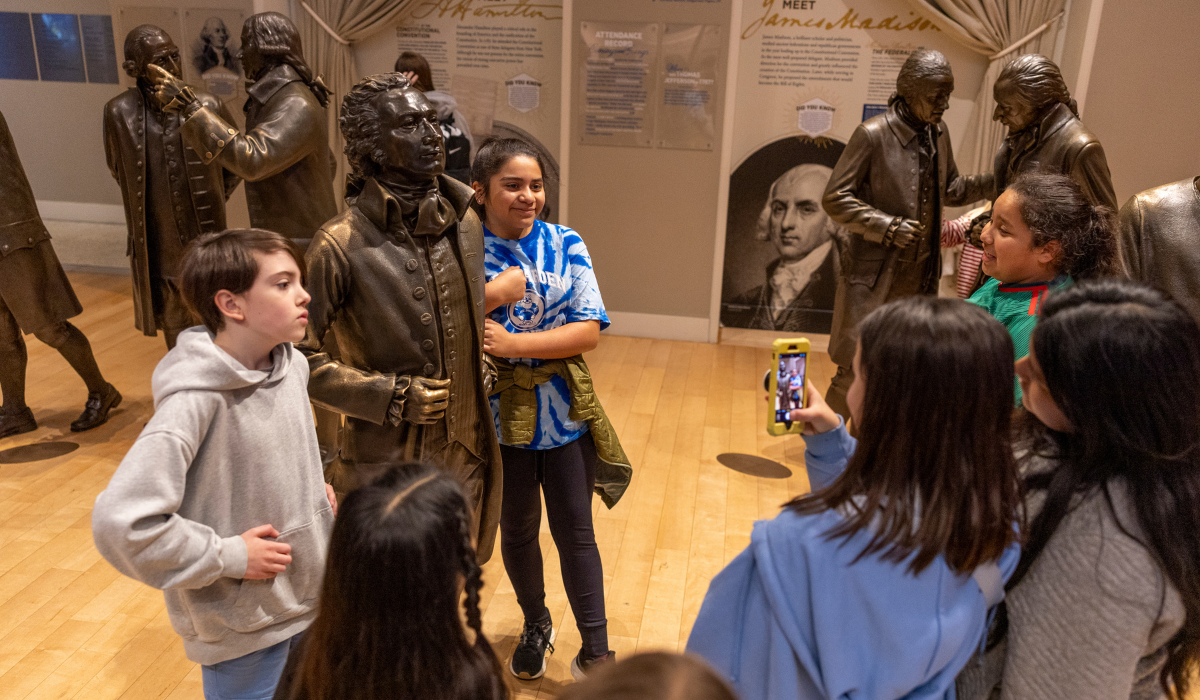 A group of students posing for photos with life-sized bronze statues of the Founding Fathers in a museum exhibit